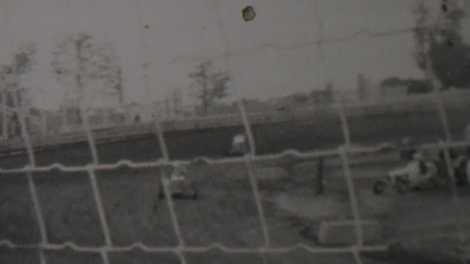 A small racing car appears behind protective netting on an outdoor dirt track in archival 1937 United States home-movie footage.