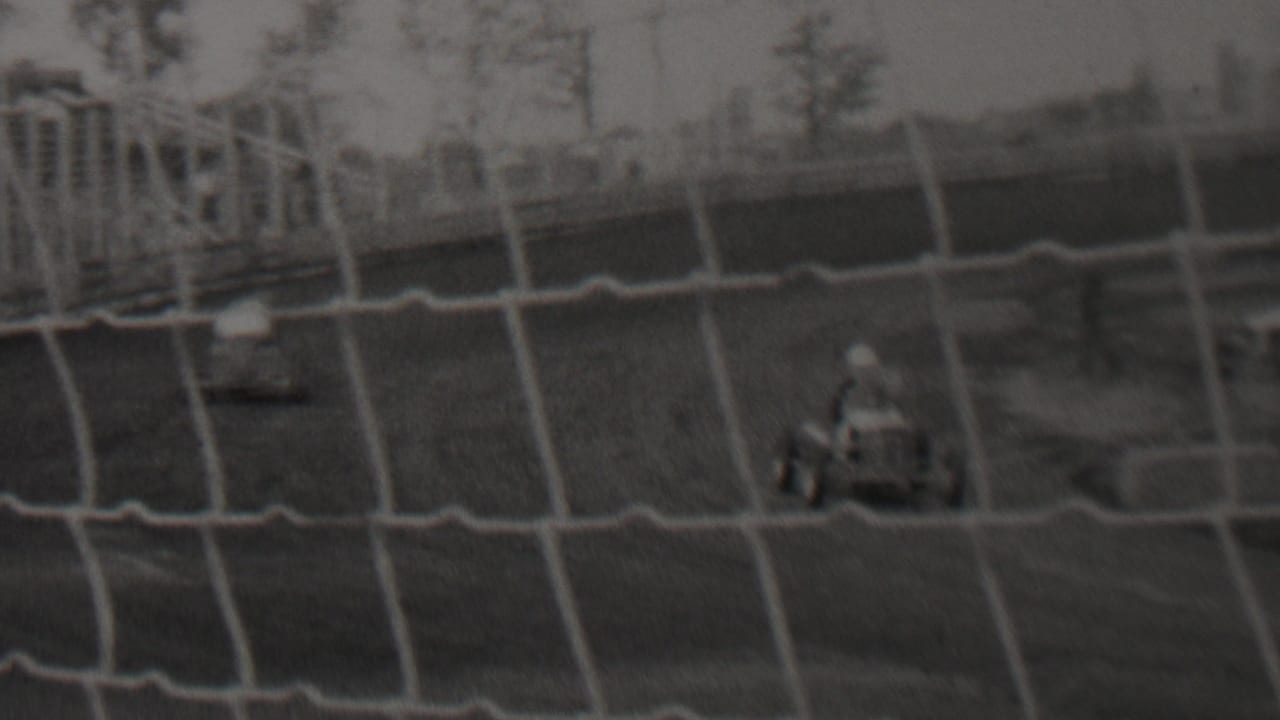 Two small cars race across a dirt track behind a net barrier.