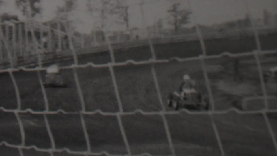 Two small cars move across a dirt racing course behind safety netting, with distant figures and trees in the background.