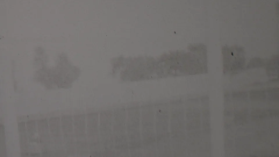The frame shows an indistinct outdoor area in the supplied 1937 Trenton home movie, with faint buildings or fences, trees, a pale vertical band, and visible film wear.