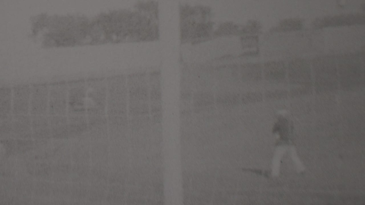 Soft black-and-white 8mm frame from Trenton, New Jersey showing a distant motorcycle rider on a dusty outdoor track with fencing and indistinct background structures.