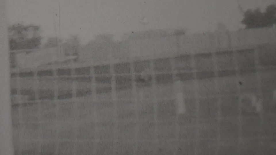 Wide black-and-white 8mm view of the race area, with fencing, low structures or barriers, trees, and open track space visible in heavy film softness.
