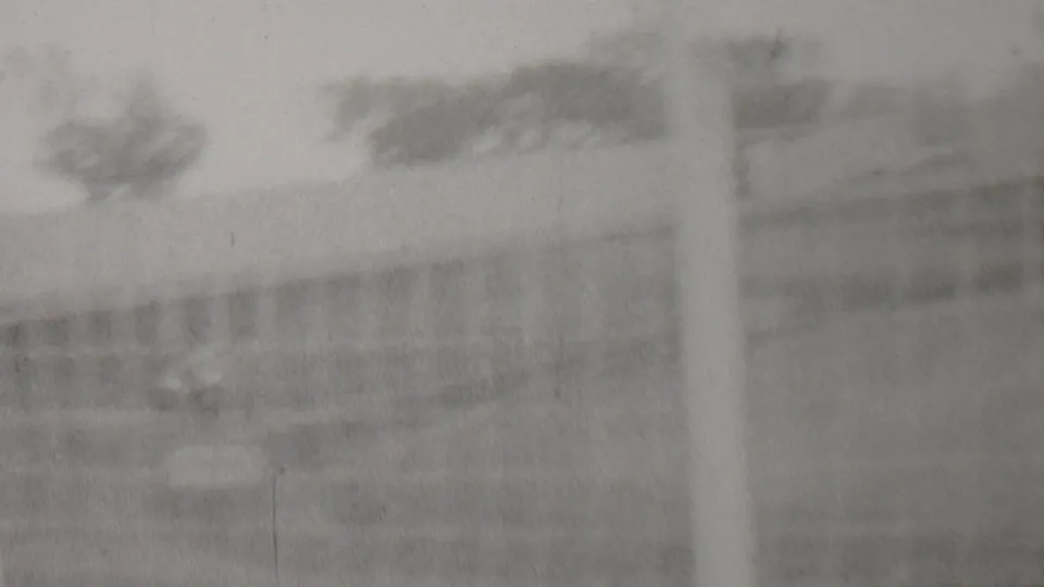 Blurred black-and-white 8mm frame showing the edge of the track area with trees, fencing or barriers, and a prominent vertical pole or post in the foreground.