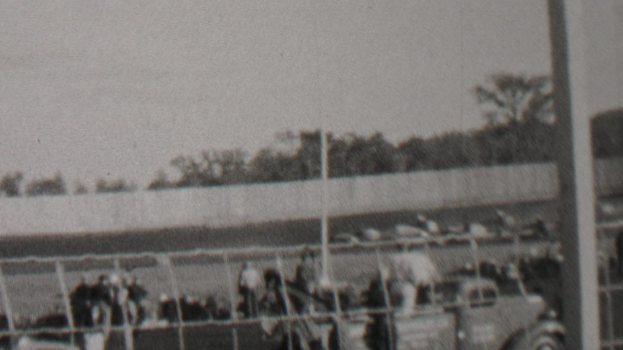 Black-and-white 8mm frame from the Trenton, New Jersey race-track clip showing spectators gathered behind fencing with automobiles and trees in the background.