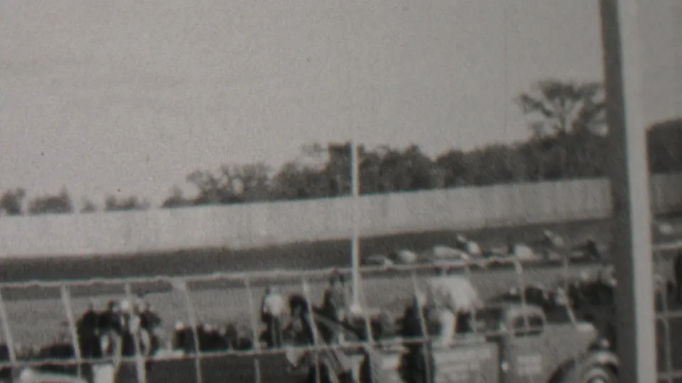 Black-and-white 8mm frame from the Trenton, New Jersey race-track clip showing spectators gathered behind fencing with automobiles and trees in the background.
