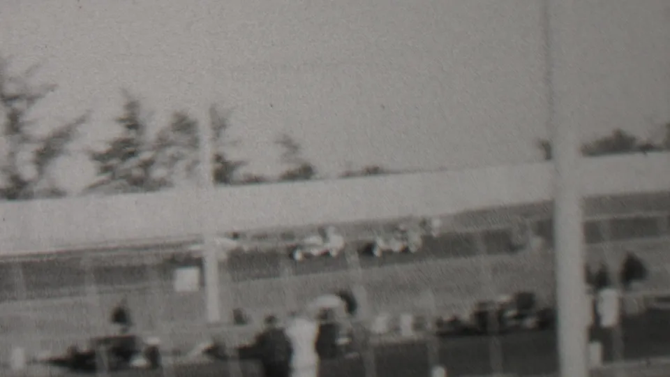 Soft black-and-white 8mm frame from the Trenton, New Jersey race-day footage showing people near the fence line with automobiles and trees behind the track.