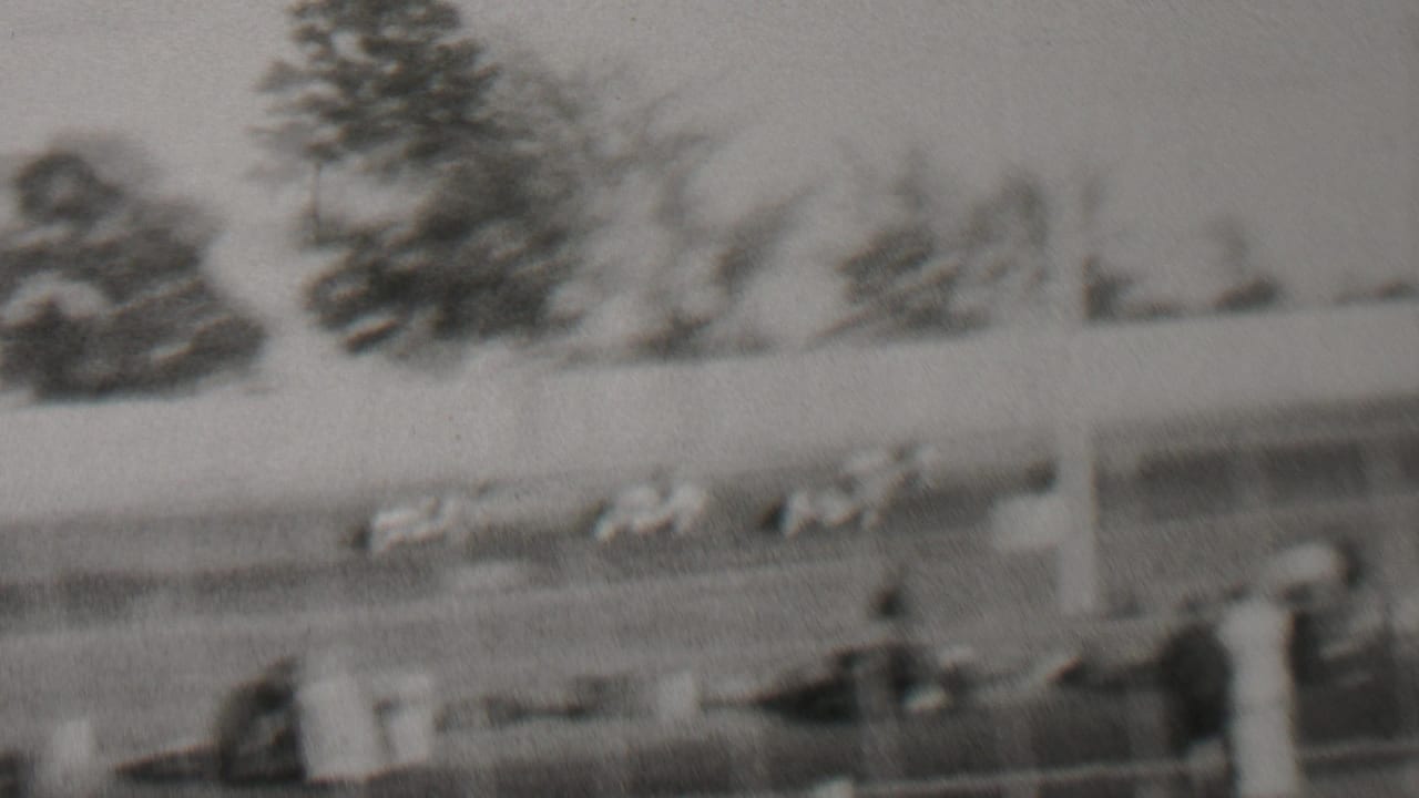 Black-and-white 1937 home-movie frame from Trenton, New Jersey showing cars on a racetrack, spectators near the fence, poles, trees, and open sky.