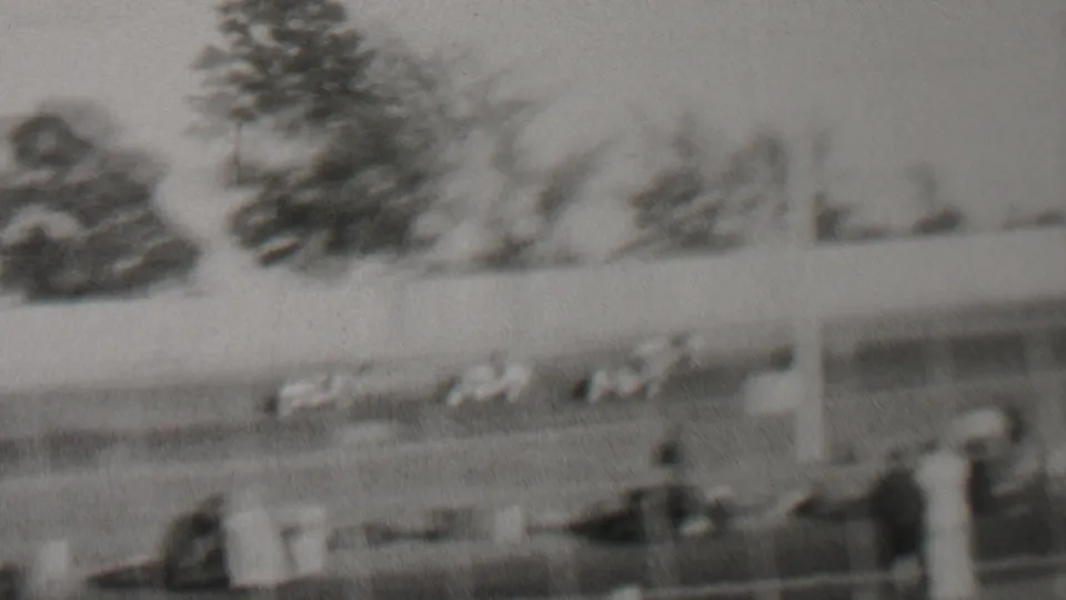 Black-and-white 1937 home-movie frame from Trenton, New Jersey showing cars on a racetrack, spectators near the fence, poles, trees, and open sky.