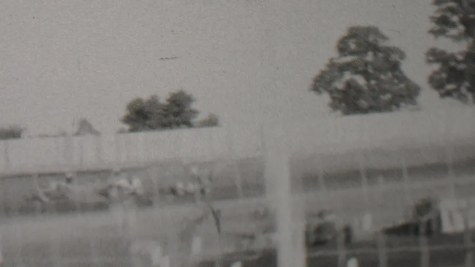 Black-and-white frame of the Trenton-area 1937 car race, with racing cars crossing the track area behind fencing and distant spectators under a bright sky.