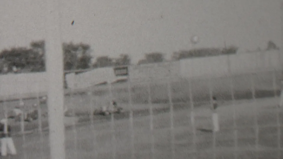 Soft black-and-white 1937 racetrack view from Trenton, New Jersey, showing fencing, a small sign, scattered spectators, cars, trees, and poles around the course.