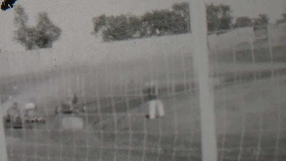 Black-and-white home-movie frame from the 1937 Trenton racing footage showing spectators by the fence, distant cars, poles, trees, and the open racetrack setting.