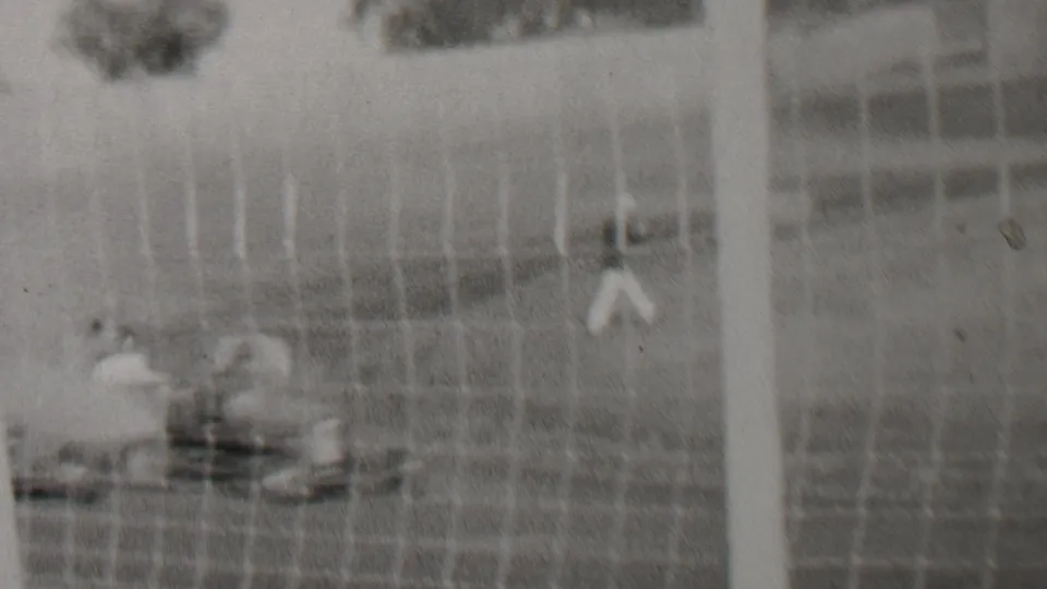1937 Trenton, New Jersey home-movie frame showing a soft black-and-white view through chain-link fencing toward a race-track area with a small distant figure and low trackside objects.