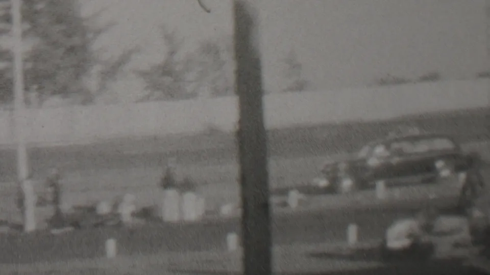 1937 Trenton, New Jersey racing footage frame with a dark vintage race car or track vehicle seen beyond foreground posts and soft fencing.