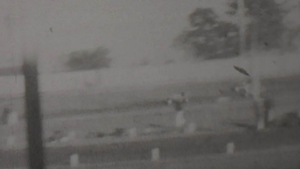 1937 Trenton, New Jersey home-movie frame showing a blurred trackside racing scene with a distant car shape, low markers, fence elements, and a person near the right edge.