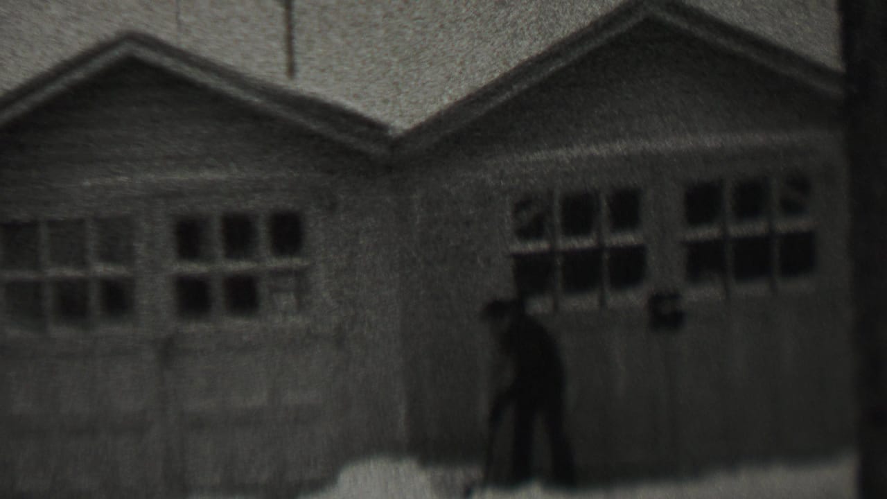 A man in dark winter clothing shovels snow near paired garage doors in 1937 Trenton, New Jersey, filmed in soft black-and-white 8mm.