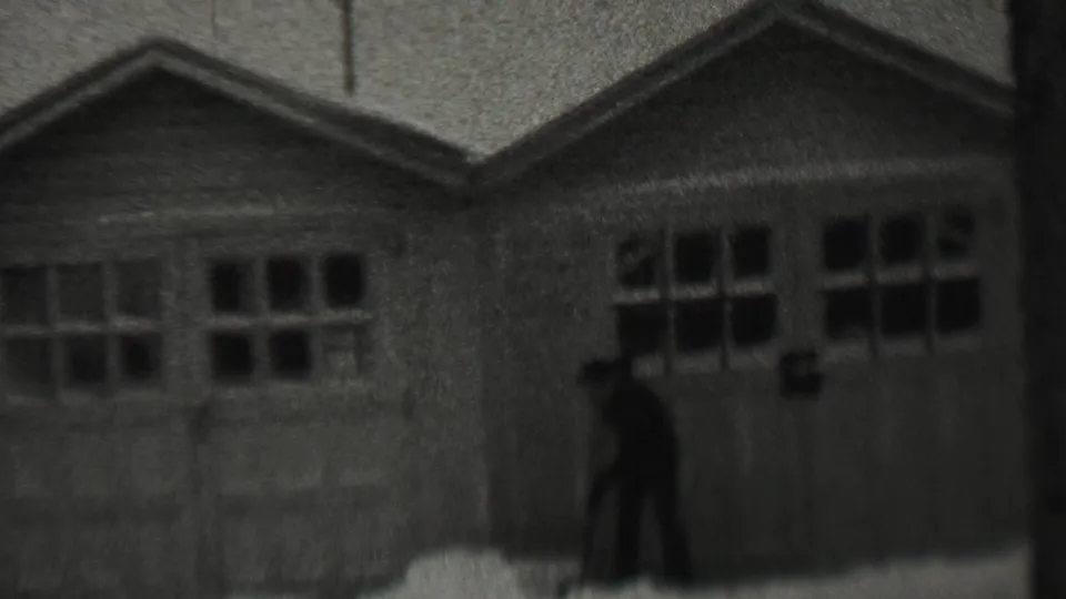 A man in dark winter clothing shovels snow near paired garage doors in 1937 Trenton, New Jersey, filmed in soft black-and-white 8mm.