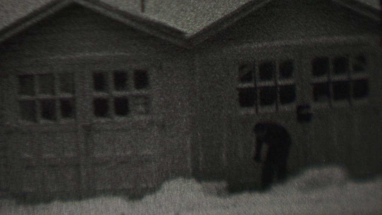 The man bends to clear snow near the garage entrance, with the residential building filling most of the frame.