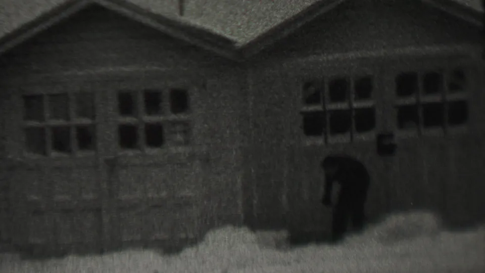 The man bends to clear snow near the garage entrance, with the residential building filling most of the frame.