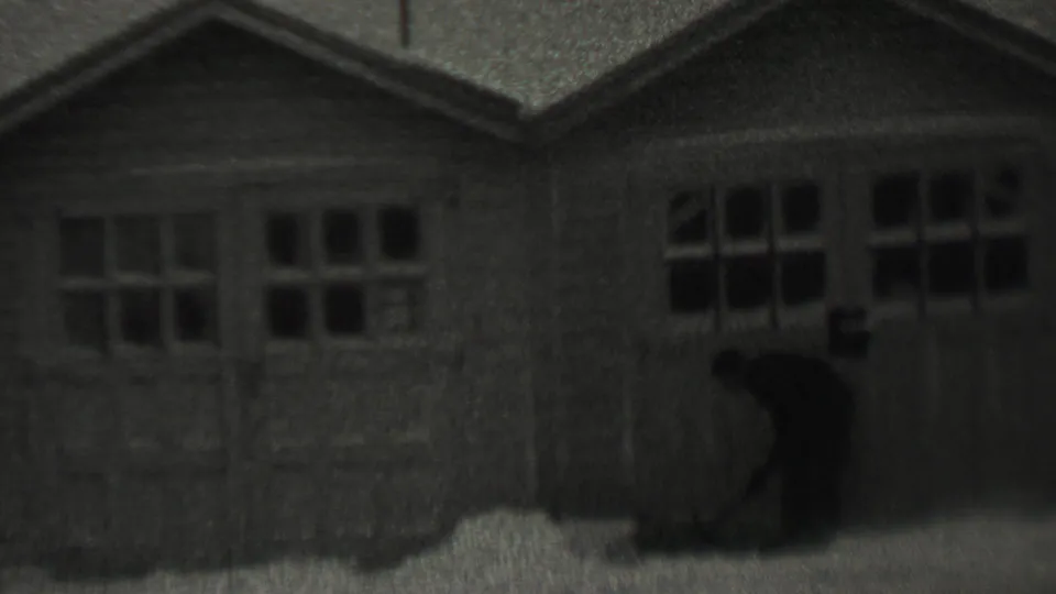 A closer view shows the man leaning into the shoveling motion in front of the garage doors, with the home-movie focus remaining soft.