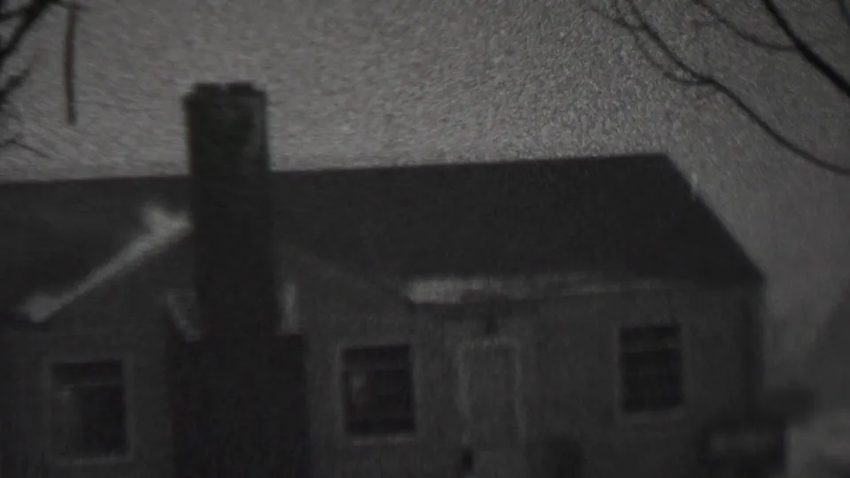 A grainy black-and-white view of the same snow-covered residential house, with bare branches crossing the upper frame.