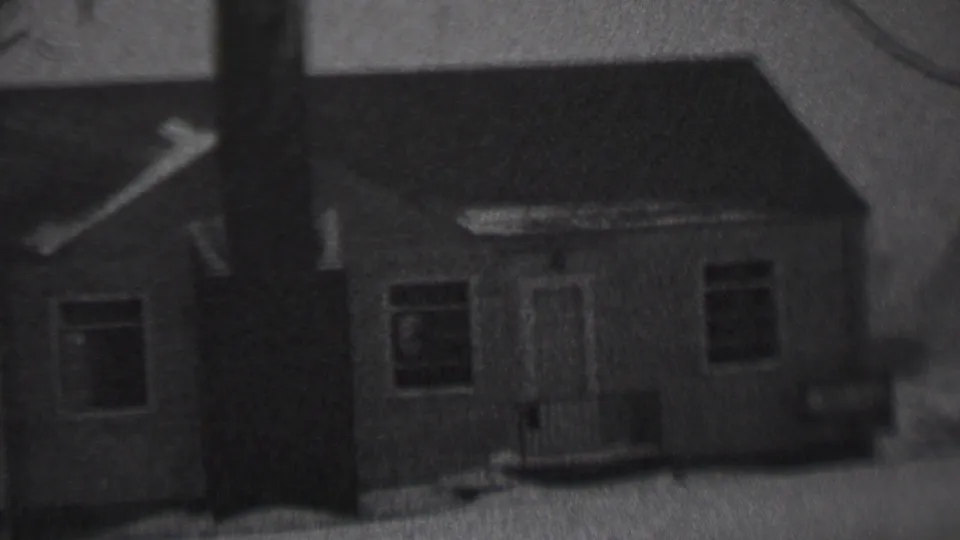 Black-and-white 8mm home-movie frame showing the front of a modest house during snowfall, with the chimney and roofline prominent.