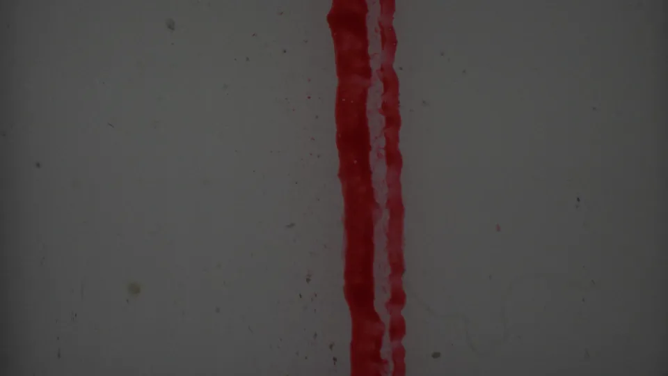 Close-up frame showing a narrow red vertical streak along the right side of a pale gray surface, with dust specks and 8mm film grain visible throughout.