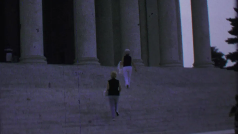 Two women in light trousers walk up broad steps beneath tall neoclassical columns in 1969 Washington DC home-movie footage.