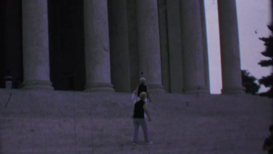 The handheld frame shows two women on a wide stairway near a columned monument, with another visitor partly visible at the edge.