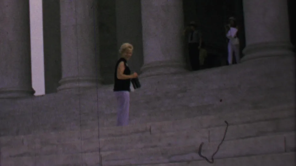 In 1969 Washington, DC home-movie footage, a woman in a sleeveless dark top and light patterned pants stands on the Lincoln Memorial steps with a camera while distant visitors remain near the columns.
