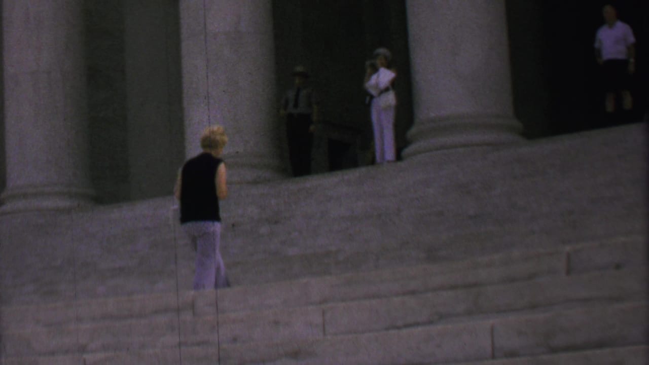 A woman in a dark sleeveless top and light patterned pants walks along the broad steps beneath the Lincoln Memorial columns, with several distant visitors in the shaded background.
