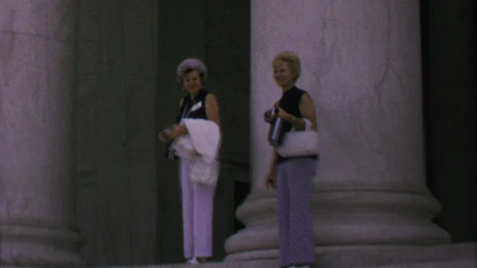 Two older women stand near a massive Lincoln Memorial column, one wearing white pants and a light hat while holding a camera and garment, the other carrying a light handbag and camera.