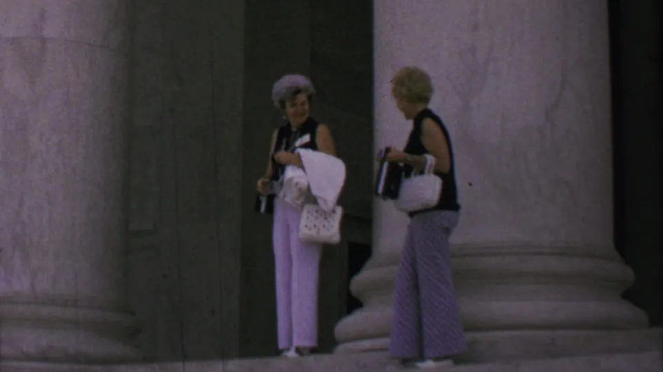 Two older women with cameras and handbags stand near the Lincoln Memorial columns, one facing the camera in white pants and a hat while the other turns toward her in patterned trousers.