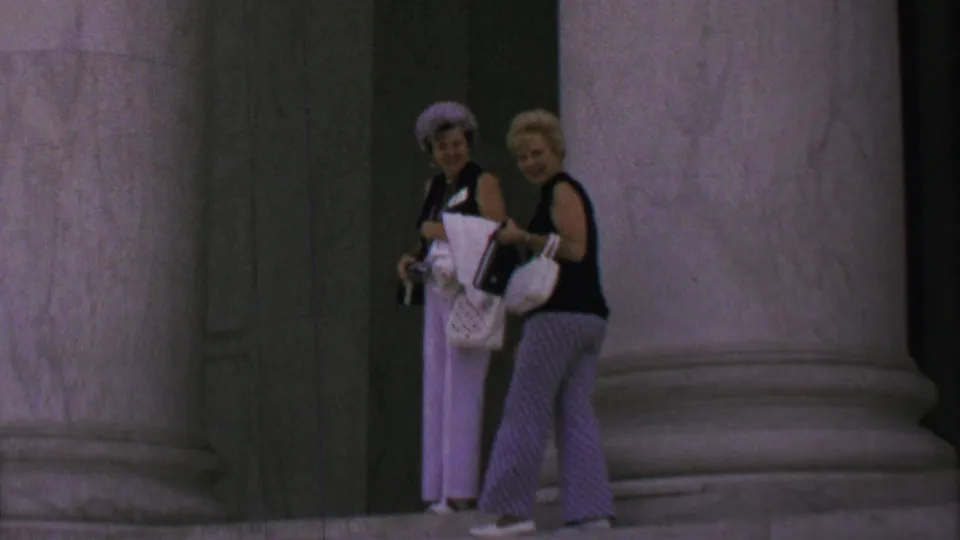 Two women in late-1960s attire stand in a Washington, DC monument colonnade, one wearing a lavender hat and light trousers while both carry white handbags.