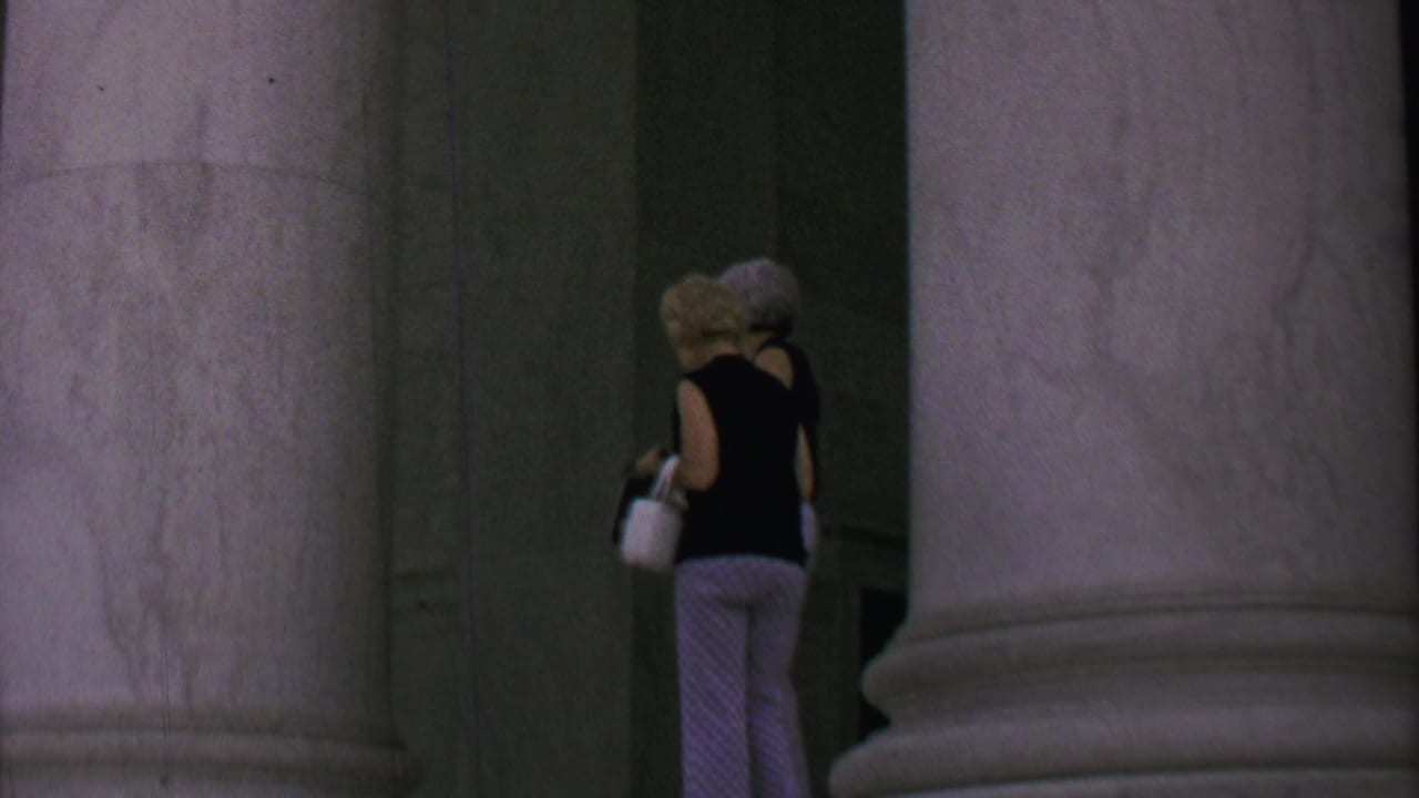 Two women walk away through a marble colonnade with large columns.
