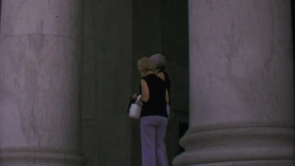 Two women with white handbags walk away through the shadowed marble columns of a Washington, DC landmark in 1969 home-movie footage.