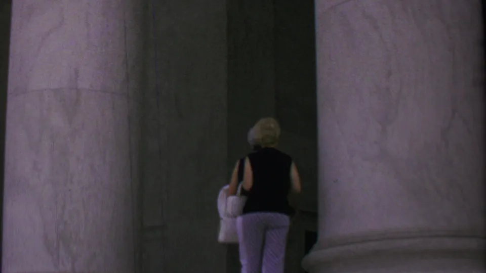 A woman in a black sleeveless top and patterned trousers carries a white handbag into a shadowed marble colonnade at a Washington, DC monument site.