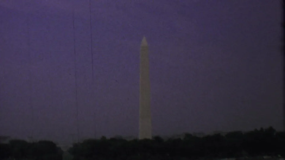 Low-light 1969 home-movie frame of the Washington Monument above a dark treeline, with purple color shift and visible film texture.