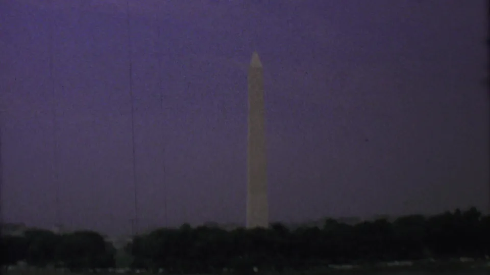 1969 8mm home-movie frame of the Washington Monument in Washington, D.C., viewed from across water with a silhouetted tree line at dusk.
