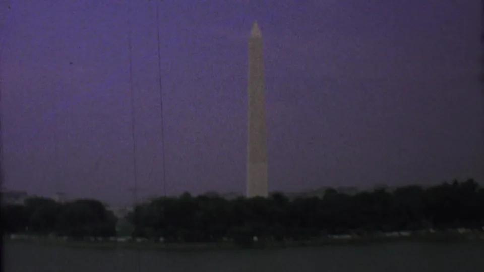 Distant 1969 home-movie view of the Washington Monument rising above a dark tree line beside water beneath a purple twilight sky.