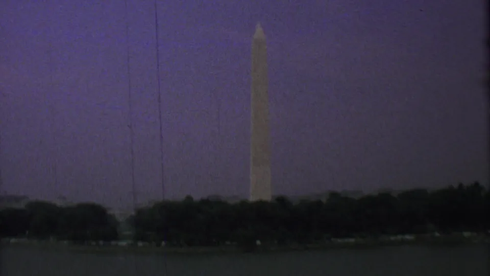 Low-light 8mm frame showing the Washington Monument across water, with dark trees and a purple evening sky dominating the scene.