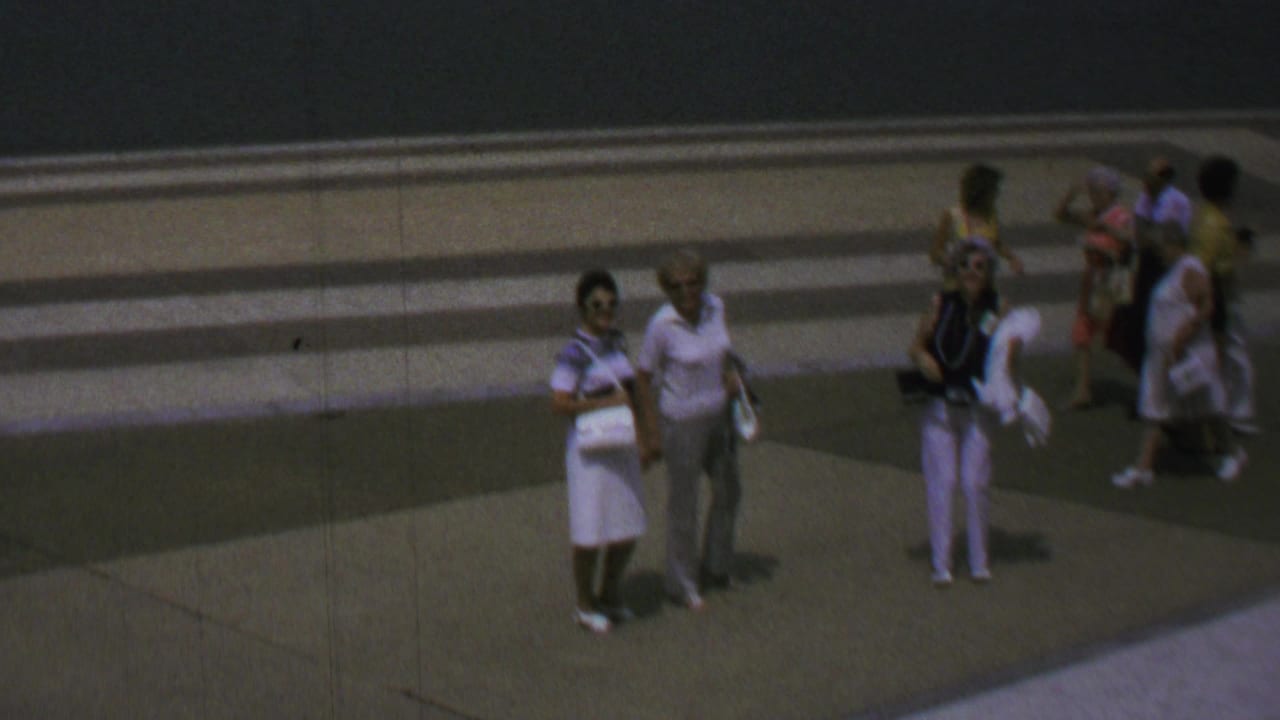 1969 Washington, DC home-movie frame with small groups of tourists crossing and pausing on a paved plaza, with period clothing visible but faces softened by distance and film grain.