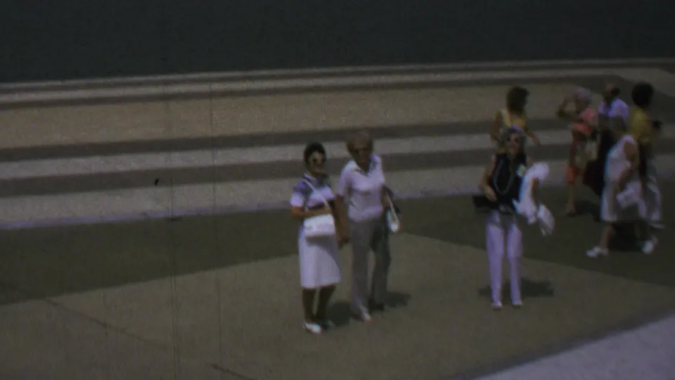 1969 Washington, DC home-movie frame with small groups of tourists crossing and pausing on a paved plaza, with period clothing visible but faces softened by distance and film grain.