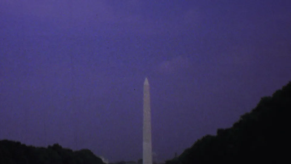 Distant 1969 Washington, DC view of the Washington Monument framed by dark trees and a purple-blue twilight sky, with the reflecting pool area faintly visible below.