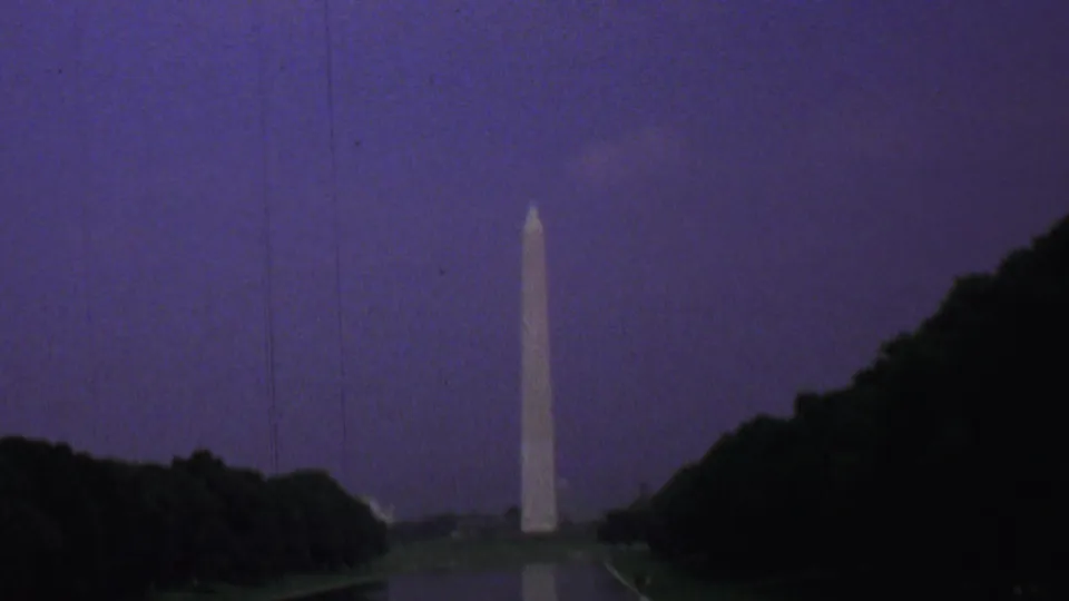 1969 Washington, DC home-movie frame showing the Washington Monument beyond the reflecting pool and tree line, with a dark foreground and strong twilight color shift.