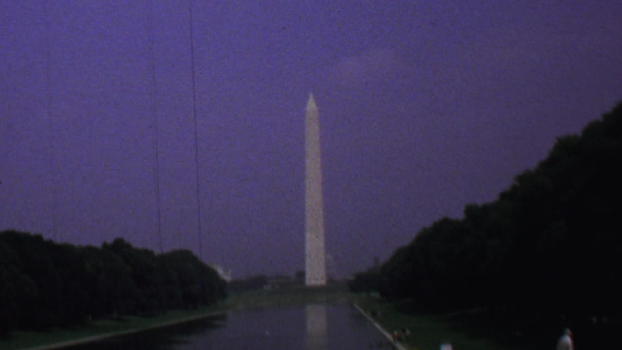 1969 8mm view of the Washington Monument seen across the Reflecting Pool in Washington DC, with dark trees framing the water under a purple dusk sky.