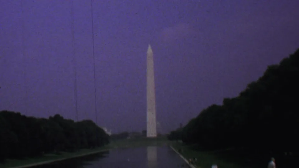 1969 8mm view of the Washington Monument seen across the Reflecting Pool in Washington DC, with dark trees framing the water under a purple dusk sky.
