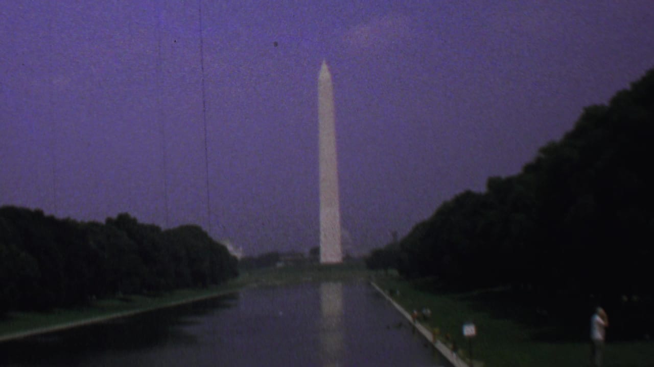 The Washington Monument stands beyond the Reflecting Pool in Washington DC, with a soft reflection in the water, dense trees along both sides, and small distant visitors on the path.