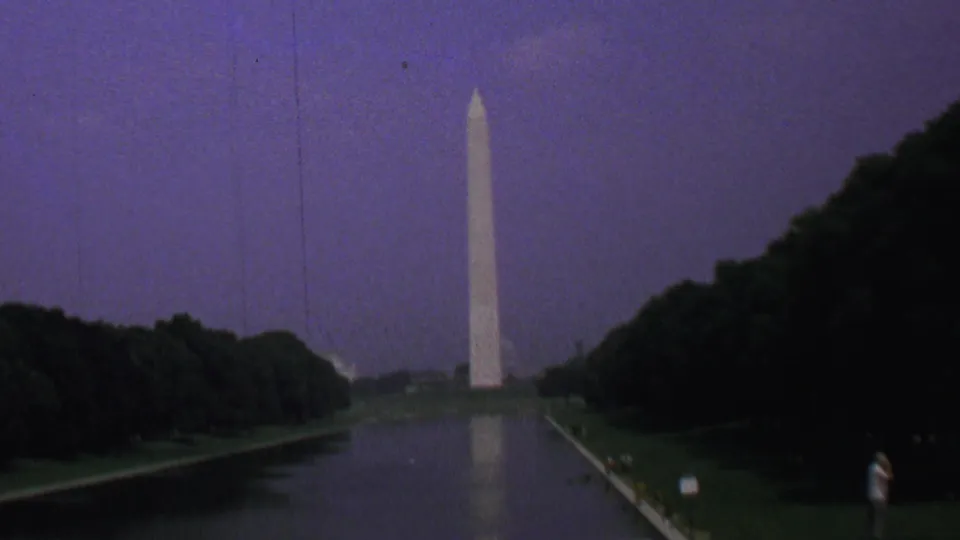 The Washington Monument stands beyond the Reflecting Pool in Washington DC, with a soft reflection in the water, dense trees along both sides, and small distant visitors on the path.