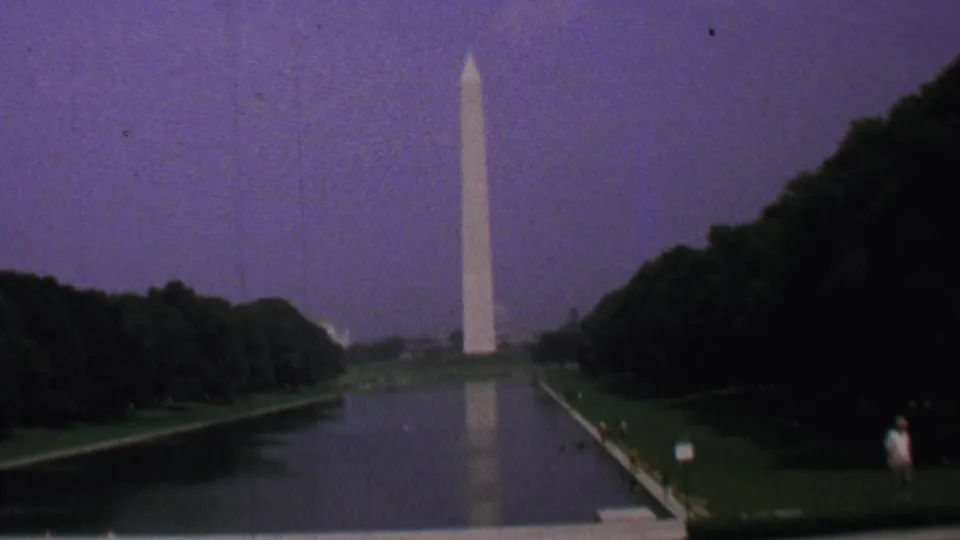 A 1969 home-movie frame looks down the Reflecting Pool toward the Washington Monument, with dark tree masses, a muted purple sky, and a small sign near the lawn.
