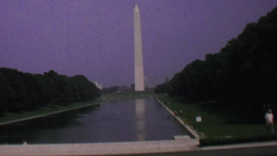 The Washington Monument rises at the far end of the Reflecting Pool in Washington DC, with trees lining the water and a distant visitor visible near the right-side path.